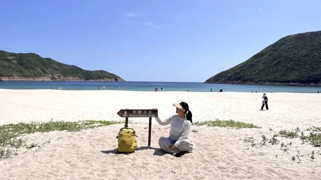 麦理浩径标牌立在沙滩上，远处是西湾碧海与绿色山峦