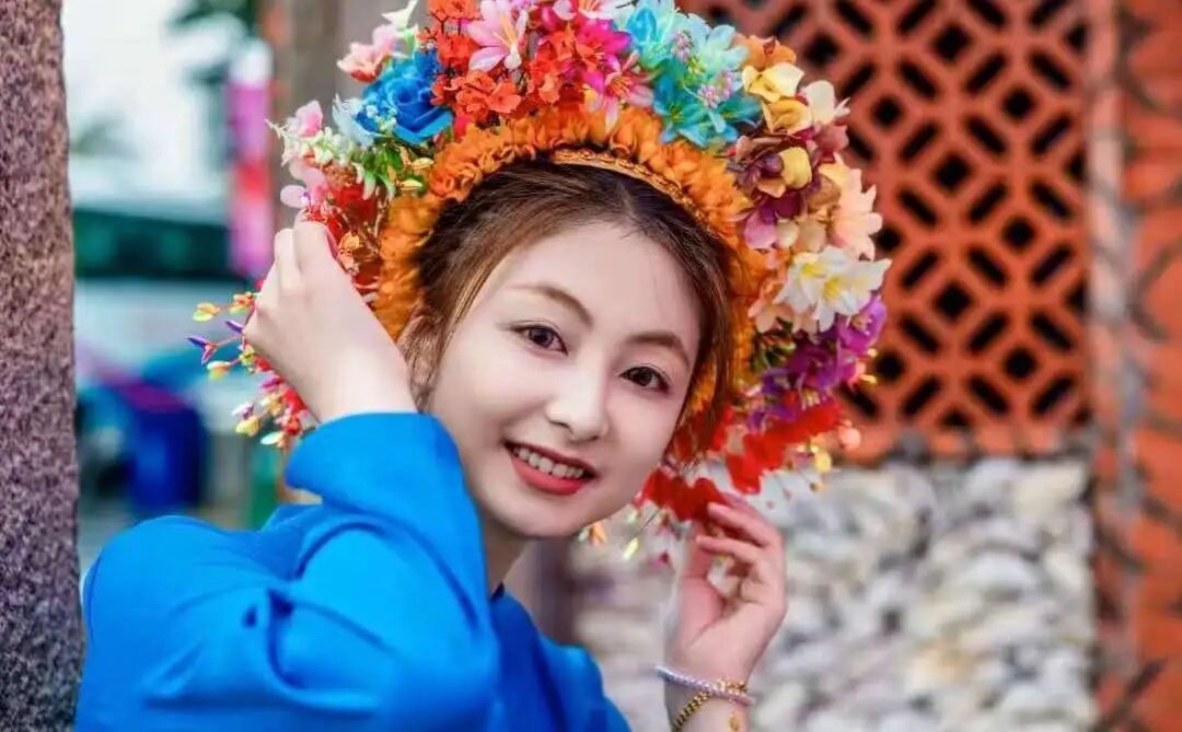 蟳埔女簪花造型的近景，头戴彩色鲜花与绢花簪成的环形花围，红砖墙作背景