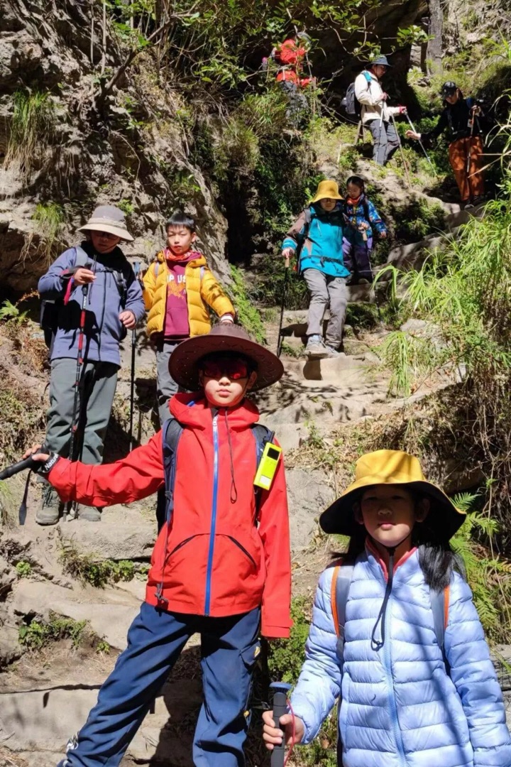 虎跳峡高路石阶上，一队亲子队员戴遮阳帽持登山杖沿峡谷崖壁向上攀登