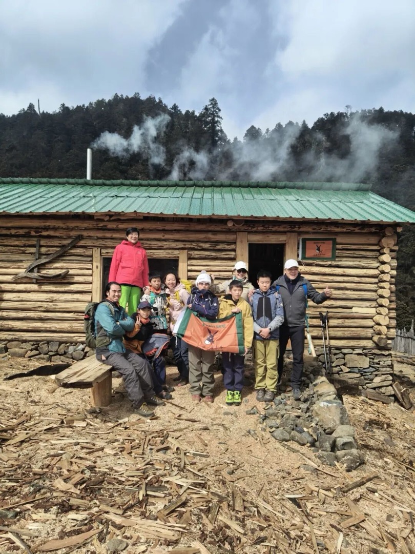 哈巴雪山云极营地木屋前，驴屋队员与孩子举着队旗合影，屋顶升起炊烟