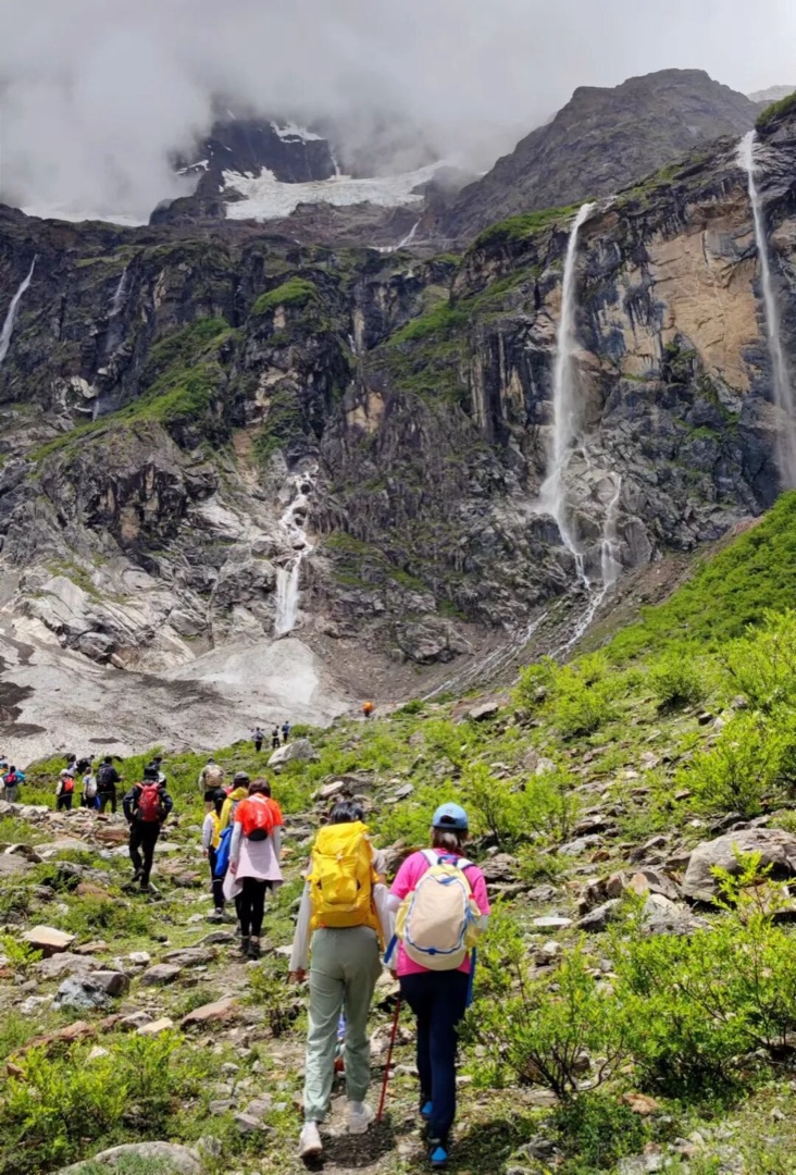 雨崩神瀑下队员背包沿碎石坡向上攀登，岩壁上多条瀑布从冰川脚下垂落